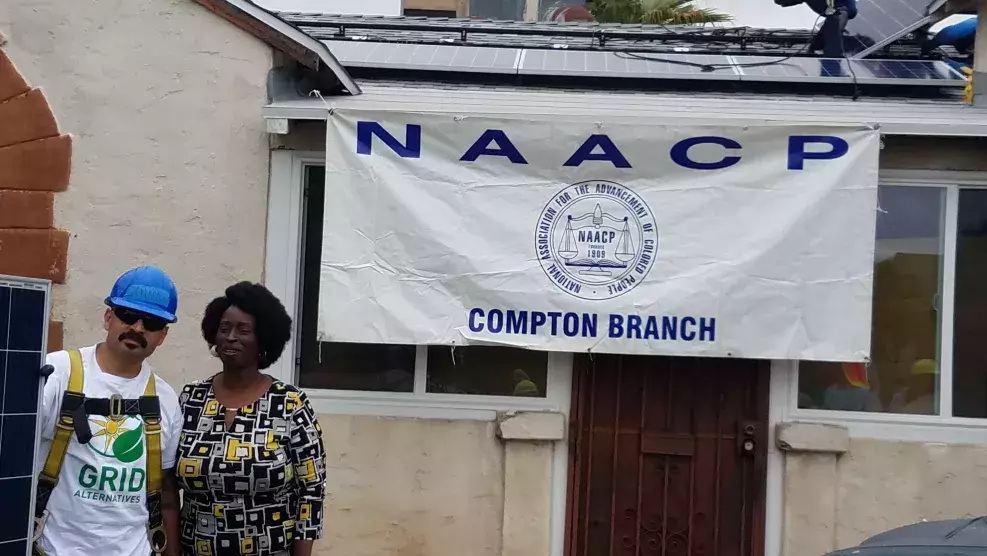 A man and woman stand side by side in front of a house on top of which an installer is putting on solar panels and a banner hangs from the roof that reads NAACP COMPTON BRANCH