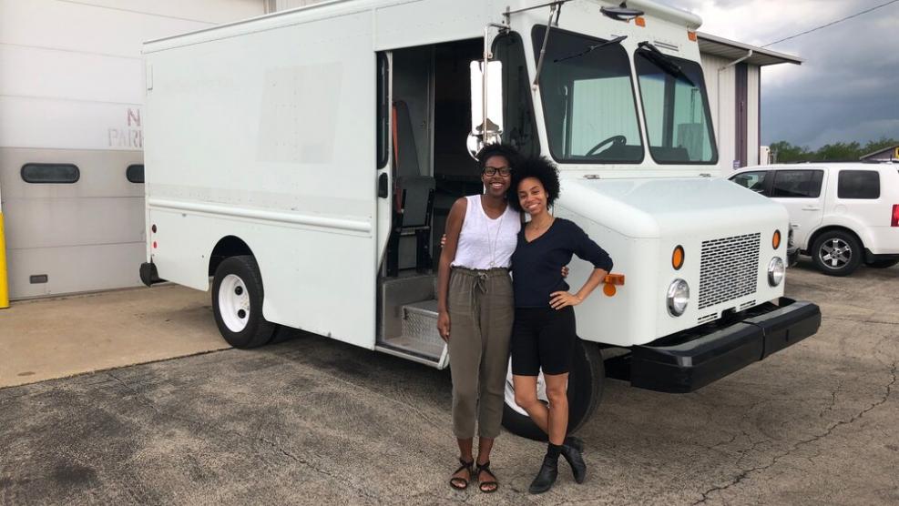 Two women embrace and smile in front of a parked white van.