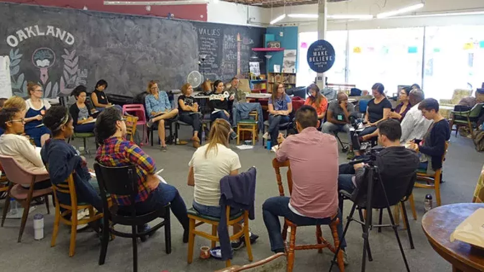 A group of people are seated in a circle in a room with a blackboard wall that reads OAKLAND.