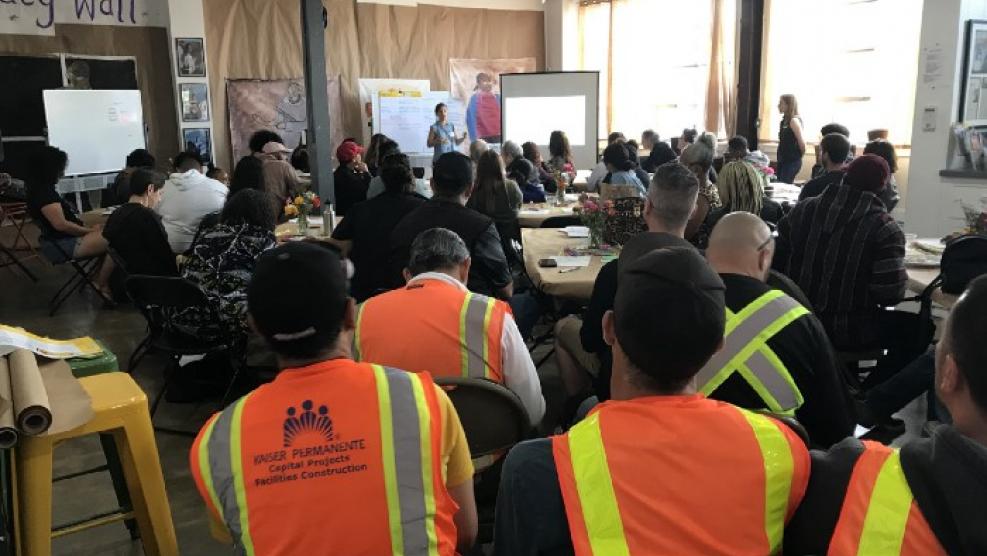 View of a room of people seated and watching a presentation, including workers in the back rows wearing orange and yellow construction vests.