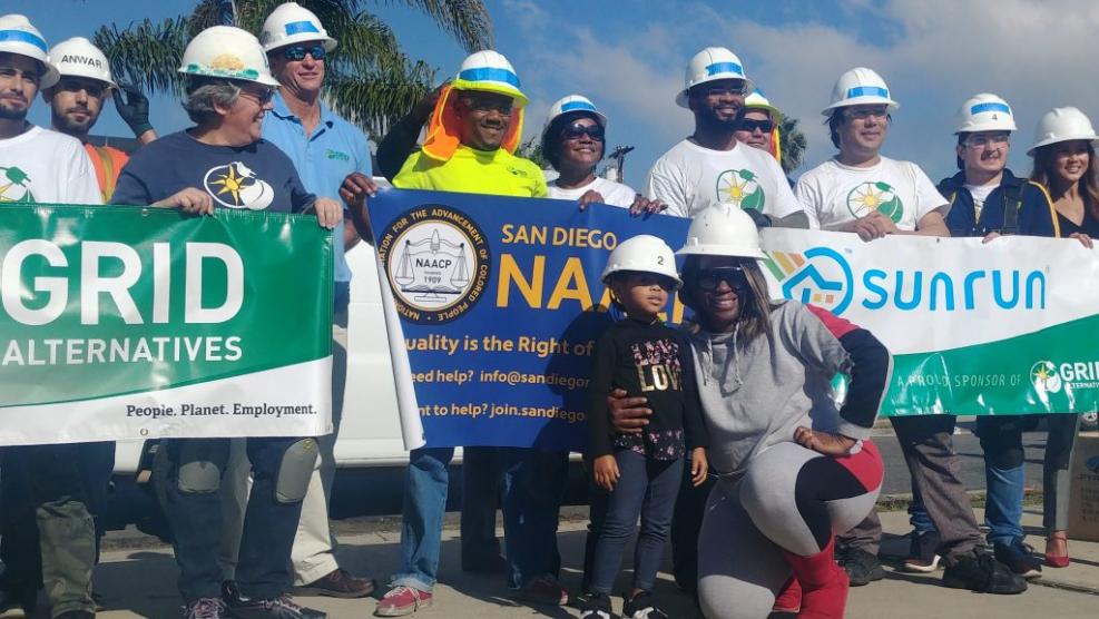 People stand in a line wearing hardhats and holding banners from GRID Alternatives, the San Diego NAACP, and Sunrun, while a woman and child kneel in front of the group.