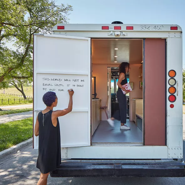 View of white van parked on a street with the back door open, a woman writing on a whiteboard, and a person standing within the van's interior.