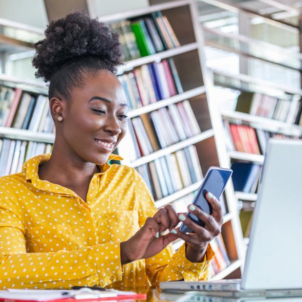 Smiling Black Woman in Library using Cell Phone and Laptop