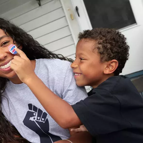 Black Child Putting "I Voted" Sticker on Woman's Cheek - Smiling