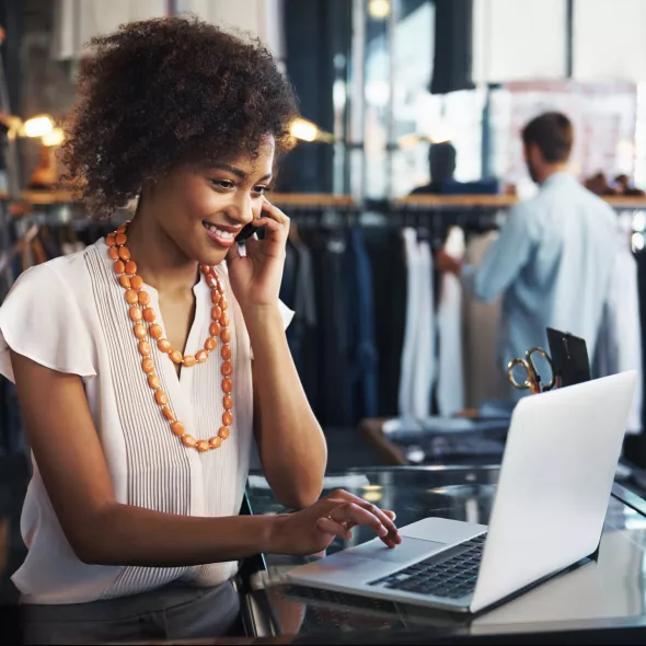 Woman Smiling and Talking into Phone and Working on Laptop in a Store Setting