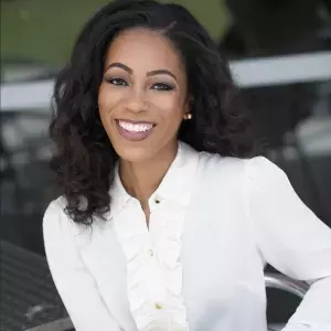 Ashley, an African American woman, smiles at the camera in a white button-down shirt. She has shoulder length dark brown hair.