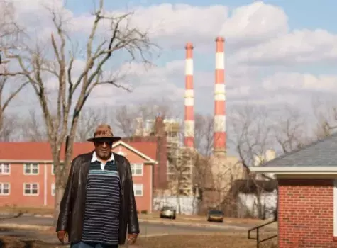 A man stands in the foreground of a neighborhood with brick multifamily buildings and smoke stacks on the horizon.