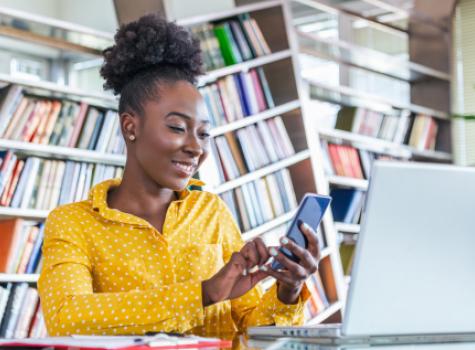 Smiling Black Woman in Library using Cell Phone and Laptop