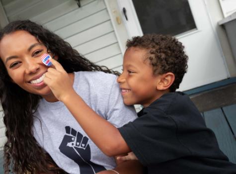 Black Child Putting "I Voted" Sticker on Woman's Cheek - Smiling